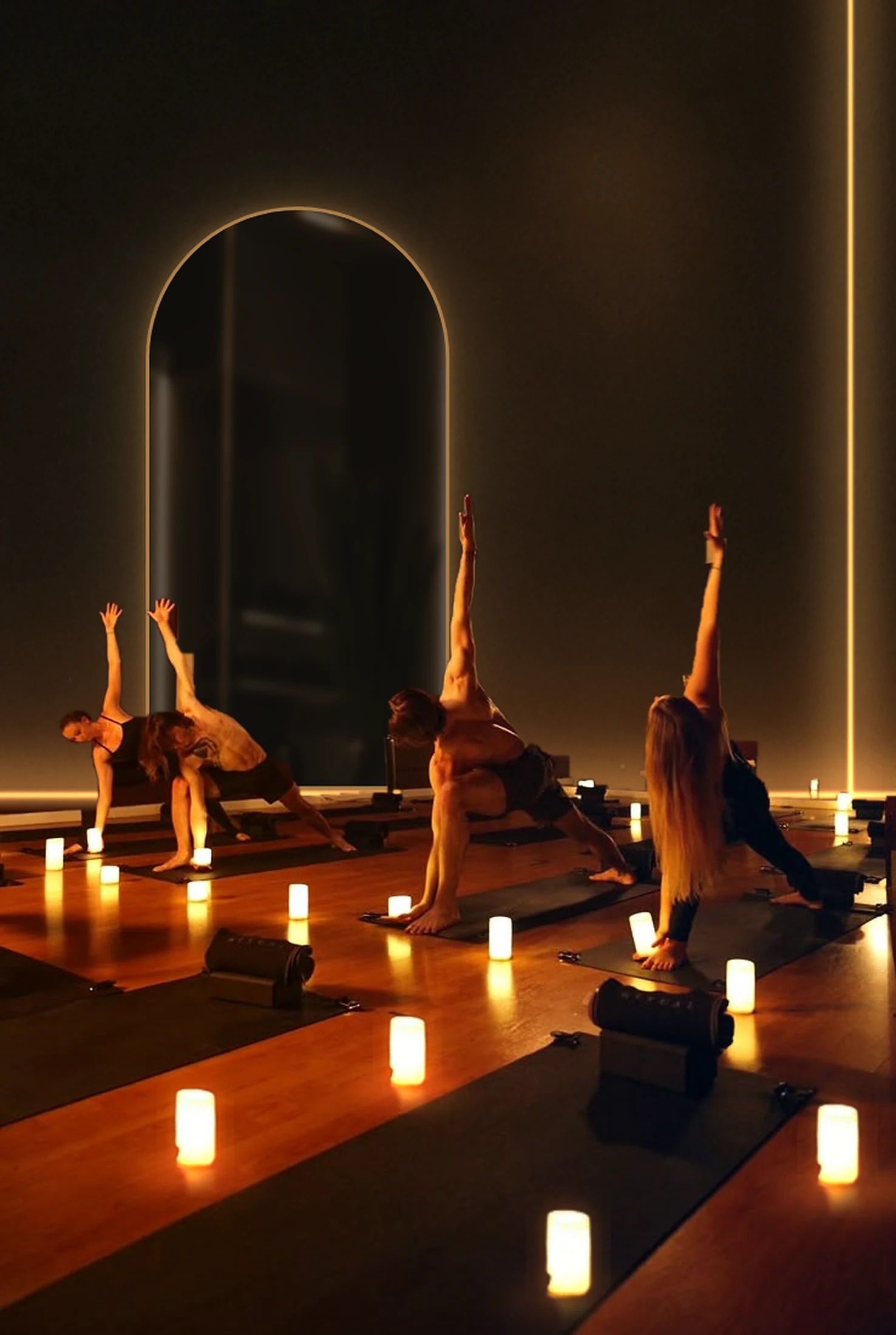 A small class holding a side-angle stretch in a candlelit studio in front of a backlit arch mirror